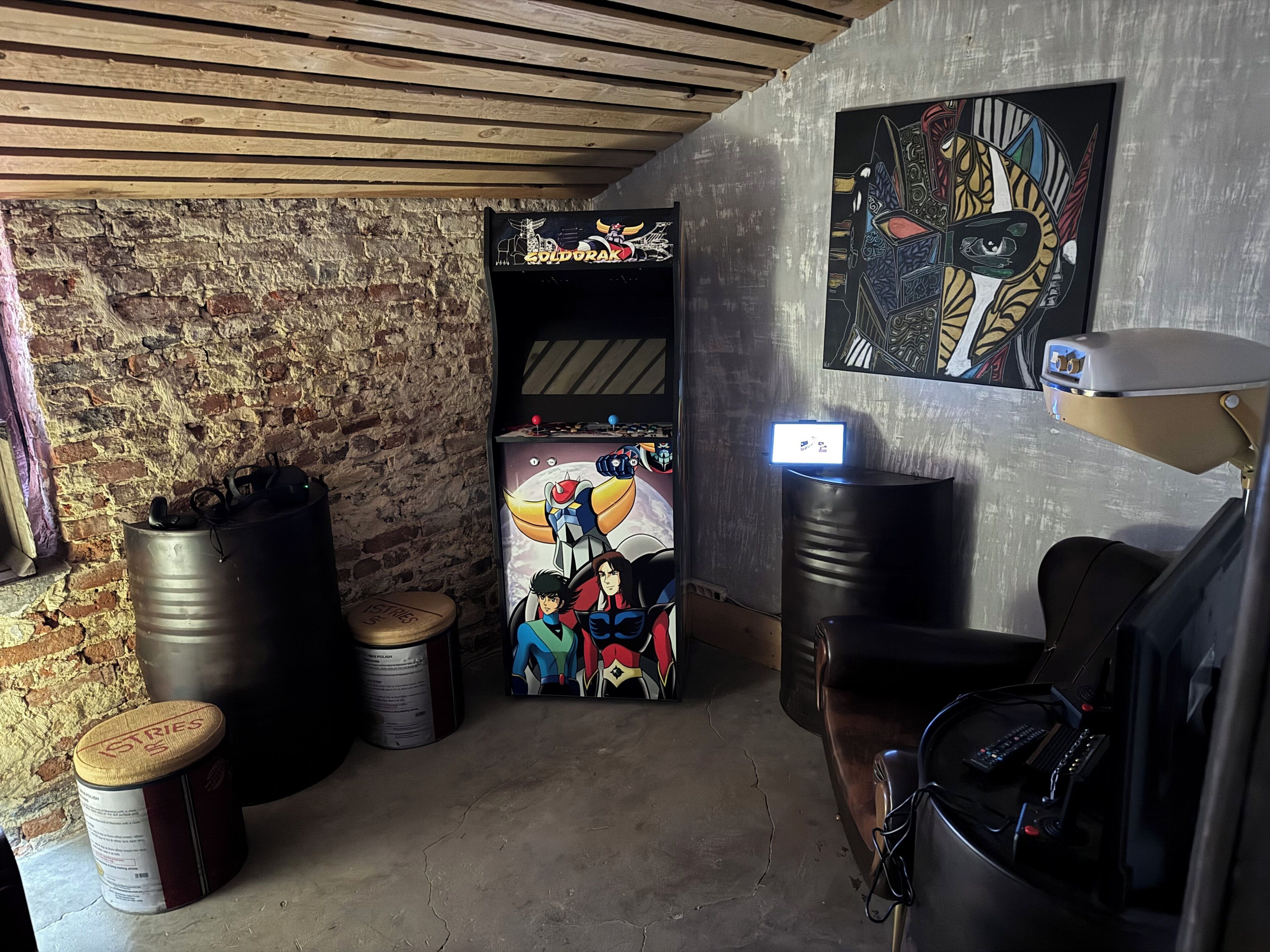 Una habitación con pared de ladrillo, techo de madera, una consola de videojuegos, un barril metálico grande, una lata de pintura y una lámpara de escritorio.