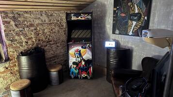 Una habitación con pared de ladrillo, techo de madera, una consola de videojuegos, un barril metálico grande, una lata de pintura y una lámpara de escritorio.