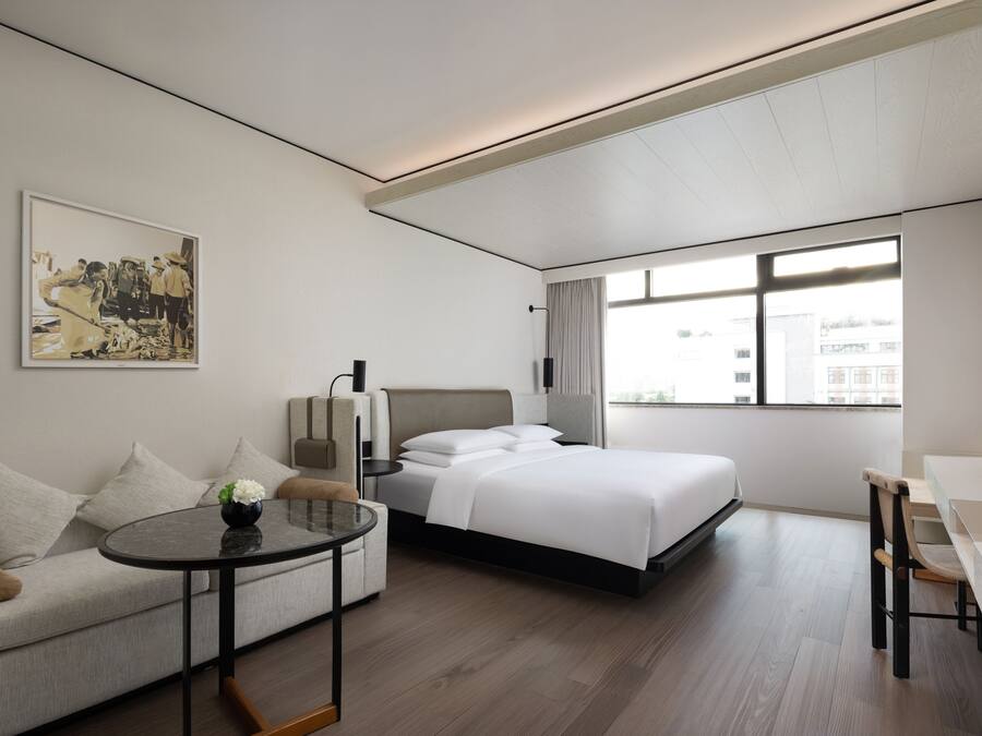 A modern hotel room with a bed, a sofa, a small table, and a large window.