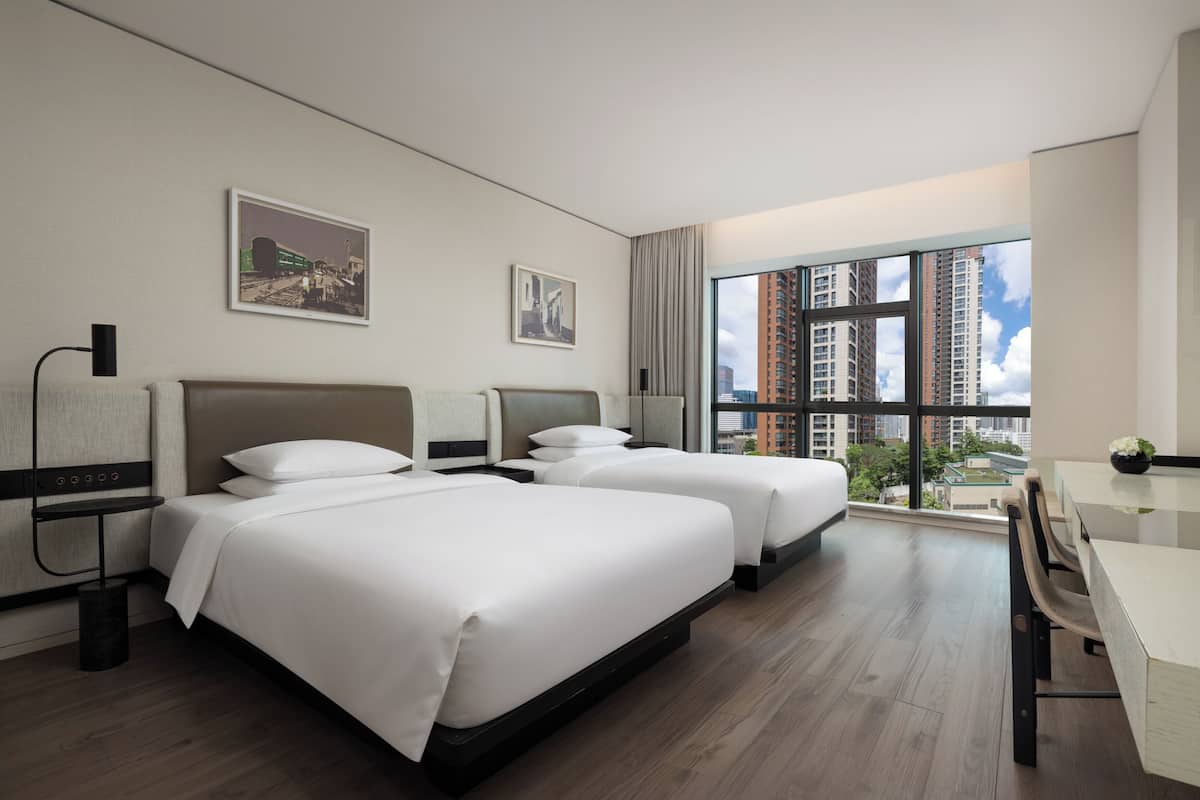 Habitación de hotel con dos camas, un escritorio y un ventanal grande con vistas a la ciudad.
