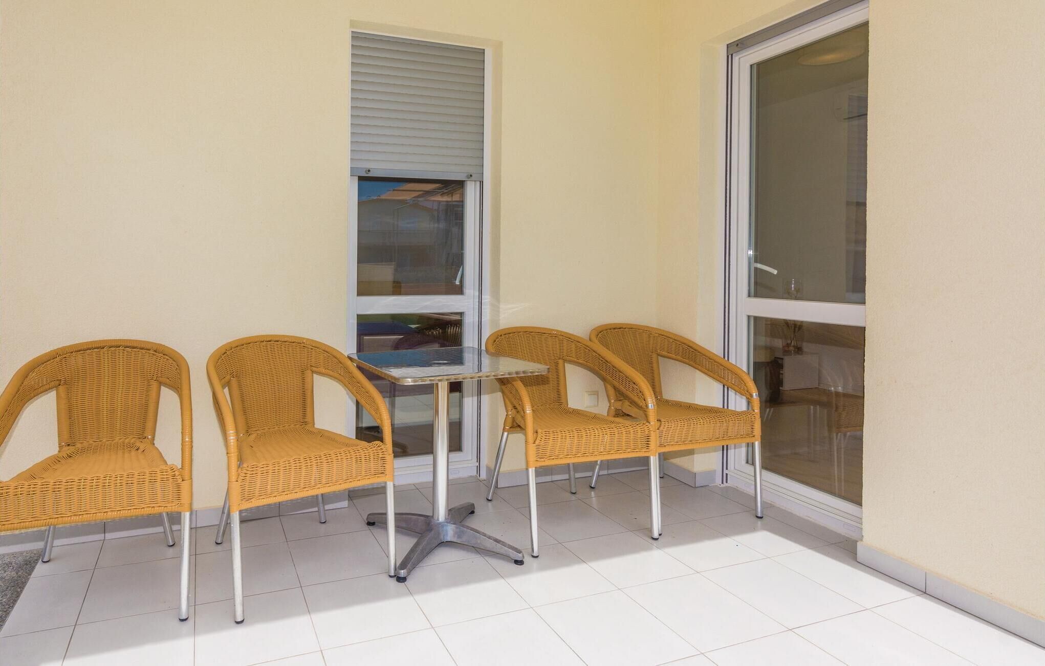 Une pièce d’angle avec quatre chaises en osier disposées autour d’une table en verre, une fenêtre avec des stores et une porte en verre.