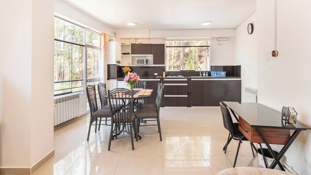 A modern kitchen with dark cabinetry, a dining area with a round table and chairs, and large windows offering a view of trees. - Amazing view in Cusco (Cusco)