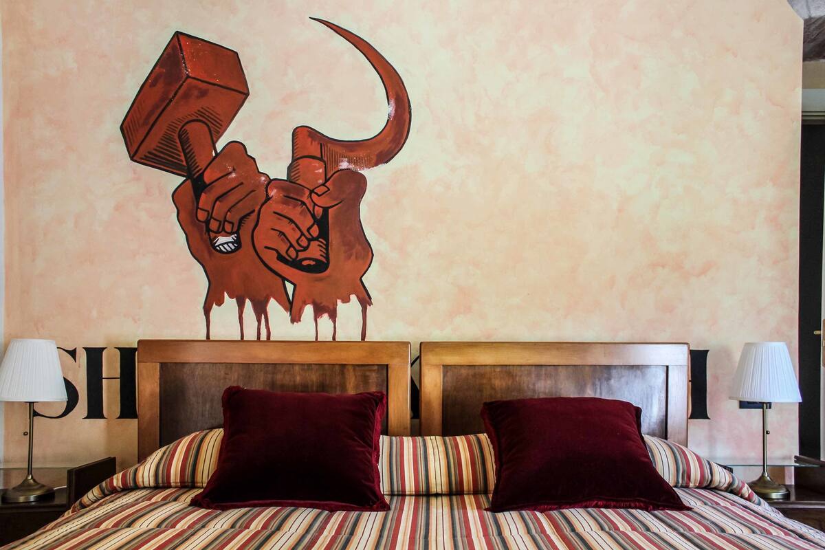 A hotel room with a bed featuring striped bedding and two red pillows, a wooden headboard, and a wall mural of a bull's head holding a hammer.