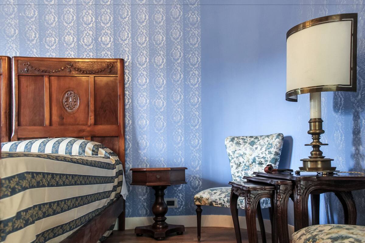 A bedroom with a wooden bed, a small wooden nightstand, a chair, and a table with a lamp.