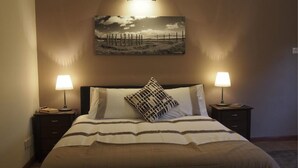 Een net opgemaakt bed met gestreept beddengoed, twee bedlampen, een aan de muur hangend schilderij en een nachtkastje van donker hout.