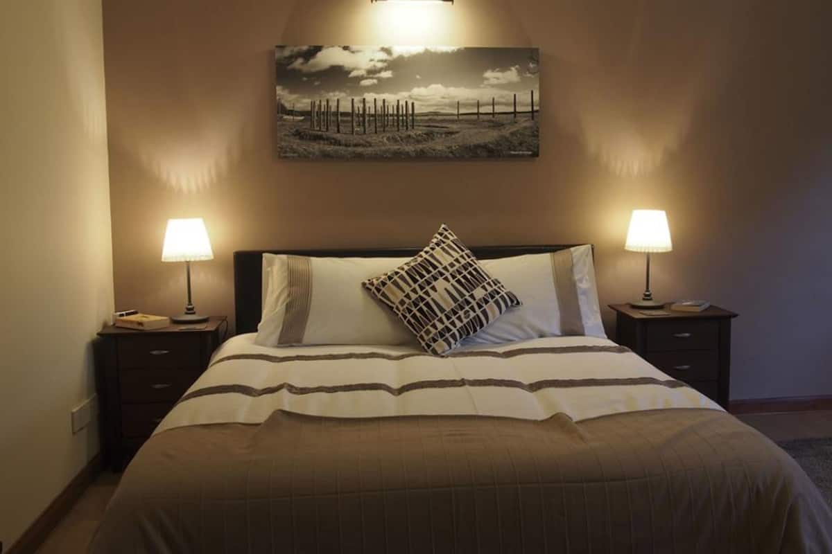 Una cama bien hecha con ropa de cama a rayas, dos lámparas de noche, un cuadro en la pared y una mesita de noche de madera oscura.