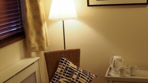 Een knus ingerichte kamer met een rieten stoel, een tafel met tijdschriften, een lamp, een lijst met een schilderij en een waterkoker op een dienblad.