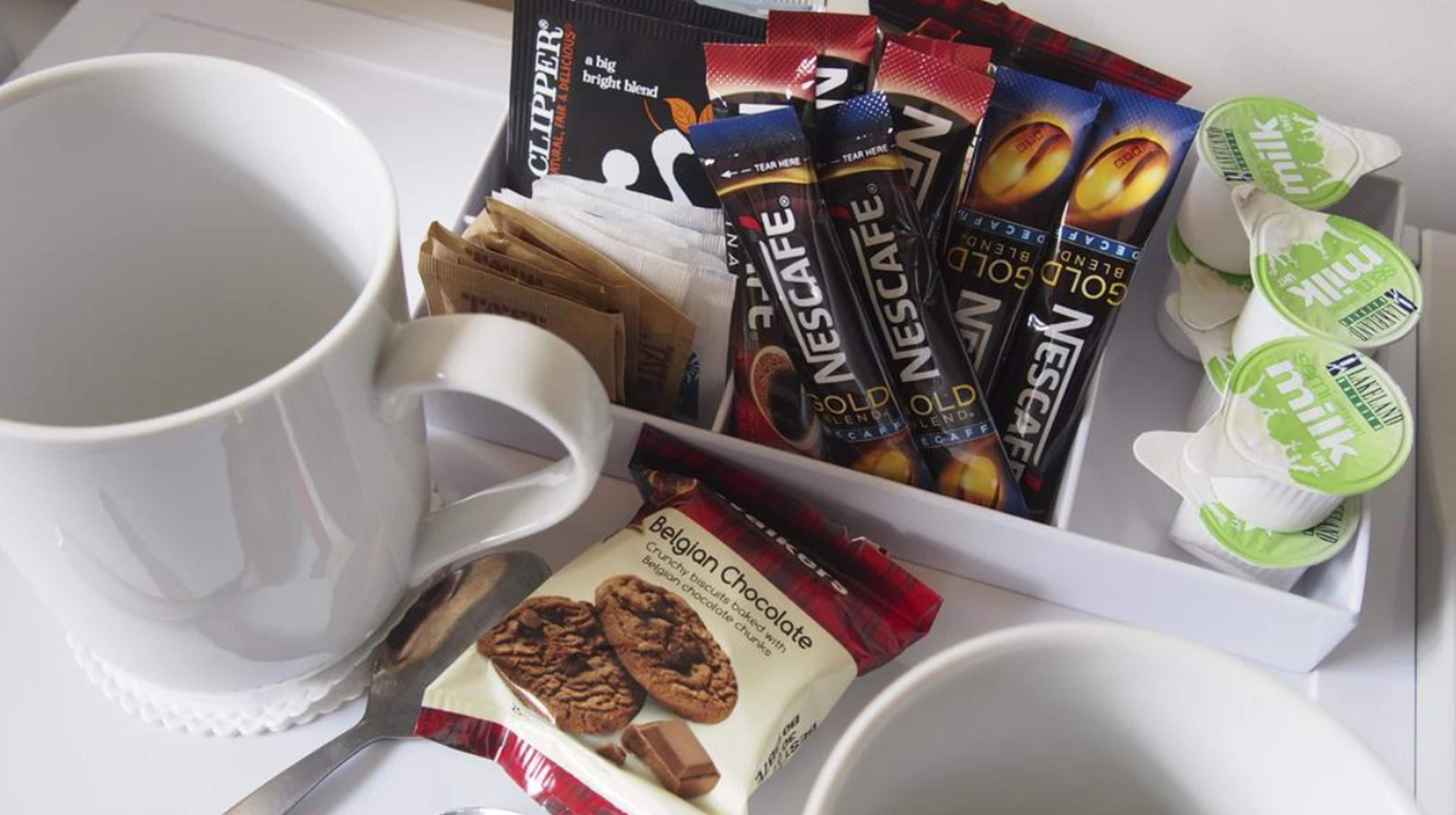 Uma bandeja com sachês de café, uma caneca, embalagem de biscoitos de chocolate belgas e um recipiente com leite.