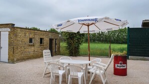 Ein Außenbereich mit weißen Plastikstühlen und einem Tisch, einem weißen Sonnenschirm mit 'Tönissteiner'-Branding und einem roten Fass mit der Aufschrift 'Jupiter'.