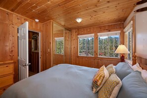 Dormitorio con paneles de madera, una cama, una mesita de noche, una lámpara y una ventana con vistas a árboles.