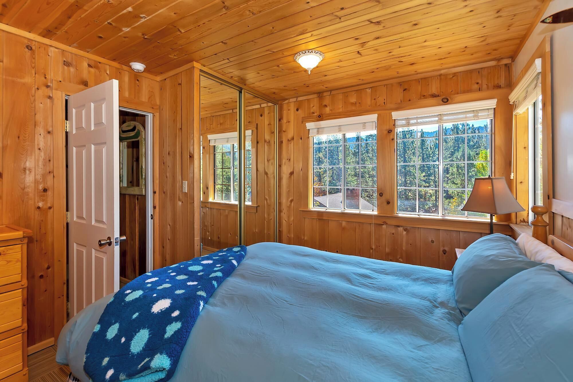 A wooden cabin bedroom with a bed, a nightstand, a lamp, and a window with a view of trees.