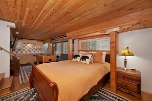 A bedroom with a wooden ceiling, a bed with a beige bedspread, a nightstand with a lamp, and a view of a kitchen and bathroom. - PET FRIENDLY!  Views of Bear Mountain Resort! Roku! Propane BBQ! Foosball! HBO Direct! Tesla Charger! (Big Bear Lake)