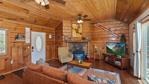 Ein gemütliches Blockhaus-Interieur mit Holzwänden, einem Kamin aus Stein, einer Couch, einem Fernseher und einem Deckenventilator.