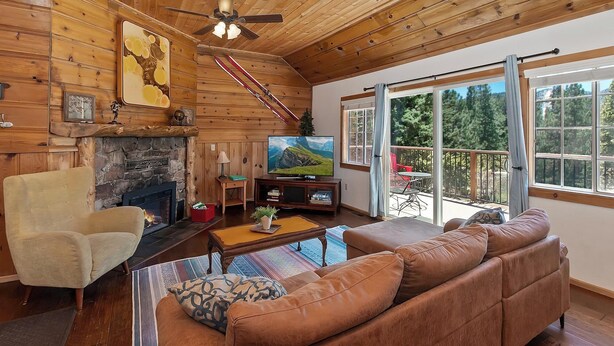 Ein gemütliches Blockhaus-Interieur mit Kamin aus Stein, einem Fernseher und einem großen Fenster mit Blick auf den Wald.