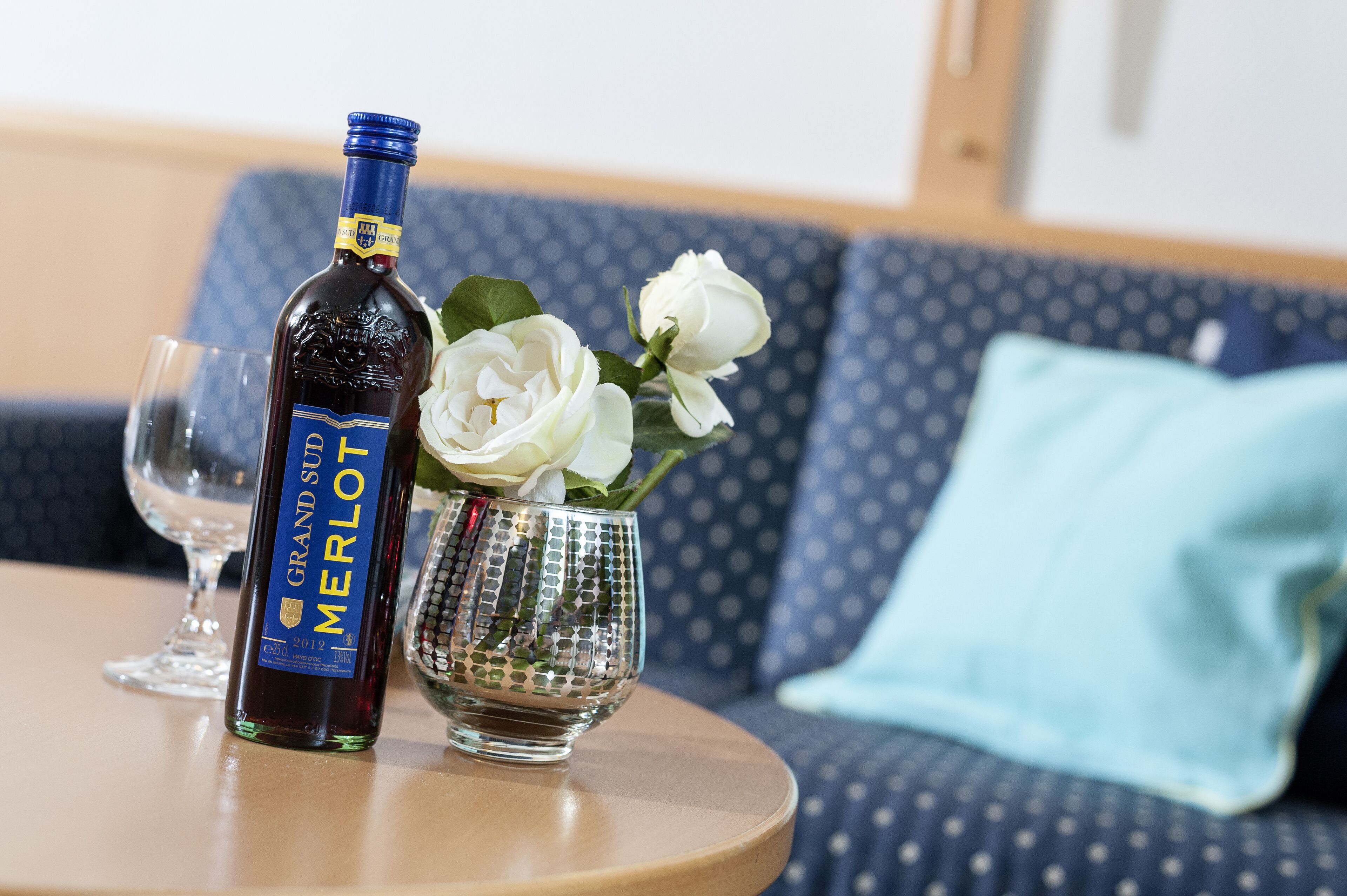 A bottle of Grand Sufi Merlot wine, a glass, and a vase with white flowers on a round table.