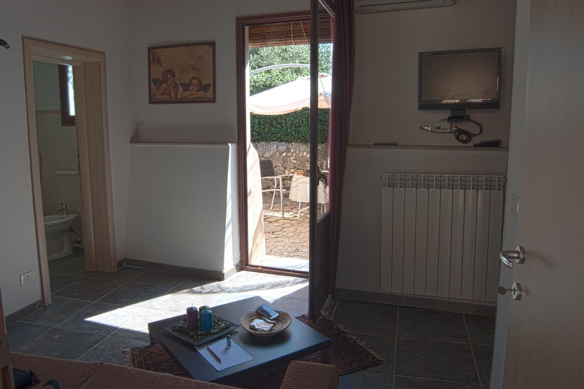 Una habitación con piso de baldosas, una pequeña mesa con una cesta y una botella, un televisor en la pared, y una puerta que da a un área exterior con patio y vistas a la vegetación.