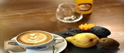 Uma chávena de café com arte de latte, um copo, uma garrafa de sumo de laranja e algumas frutas sobre uma mesa.