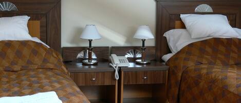 A hotel room with two beds, each with a patterned duvet cover, a desk with two lamps, a telephone, and a small shelf.