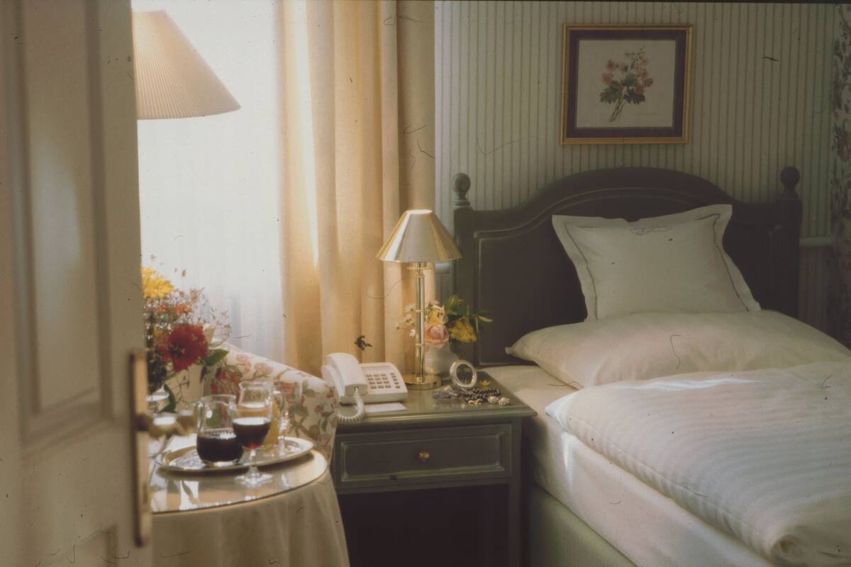 A hotel room with a bed, bedside table with a lamp, a telephone, and a vase with flowers.