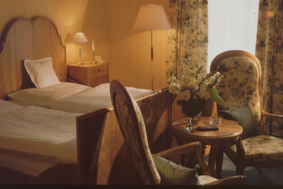 A hotel room with a bed, bedside table with a lamp, a chair, a small table with a vase of flowers, and a window with curtains.