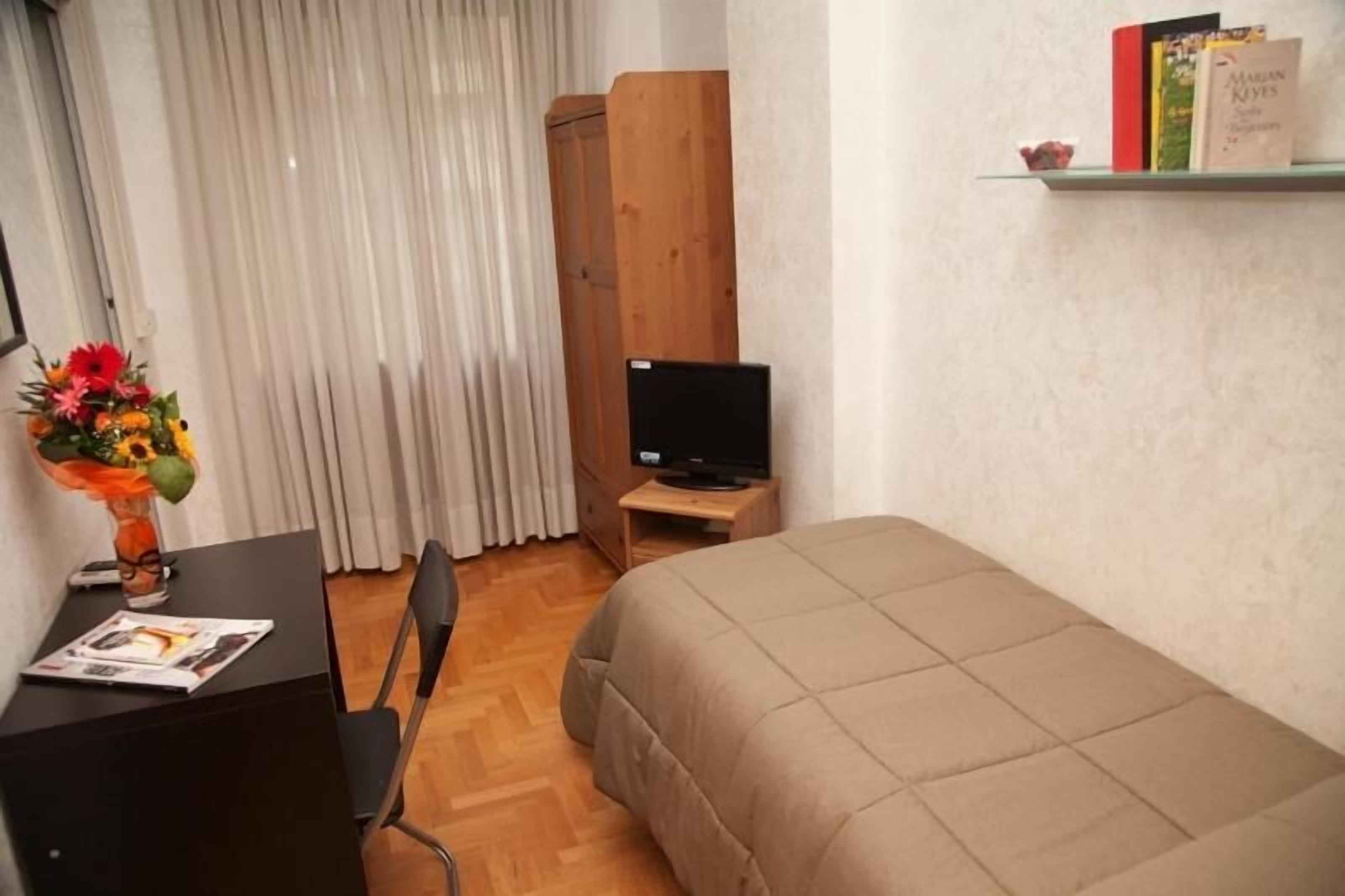 Ein Schlafzimmer mit einem Bett, einem Schreibtisch mit Stuhl, einem Fernseher auf einem Holzboden, einem Bücherregal mit Büchern und einem Fenster mit Vorhängen.