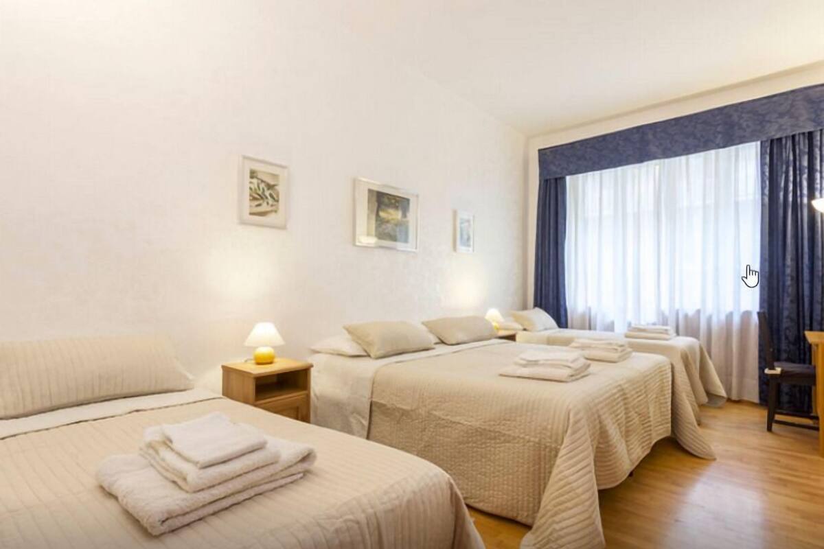 A hotel room with three beds, each with a white duvet and pillows, a bedside table with a lamp, and a window with curtains.