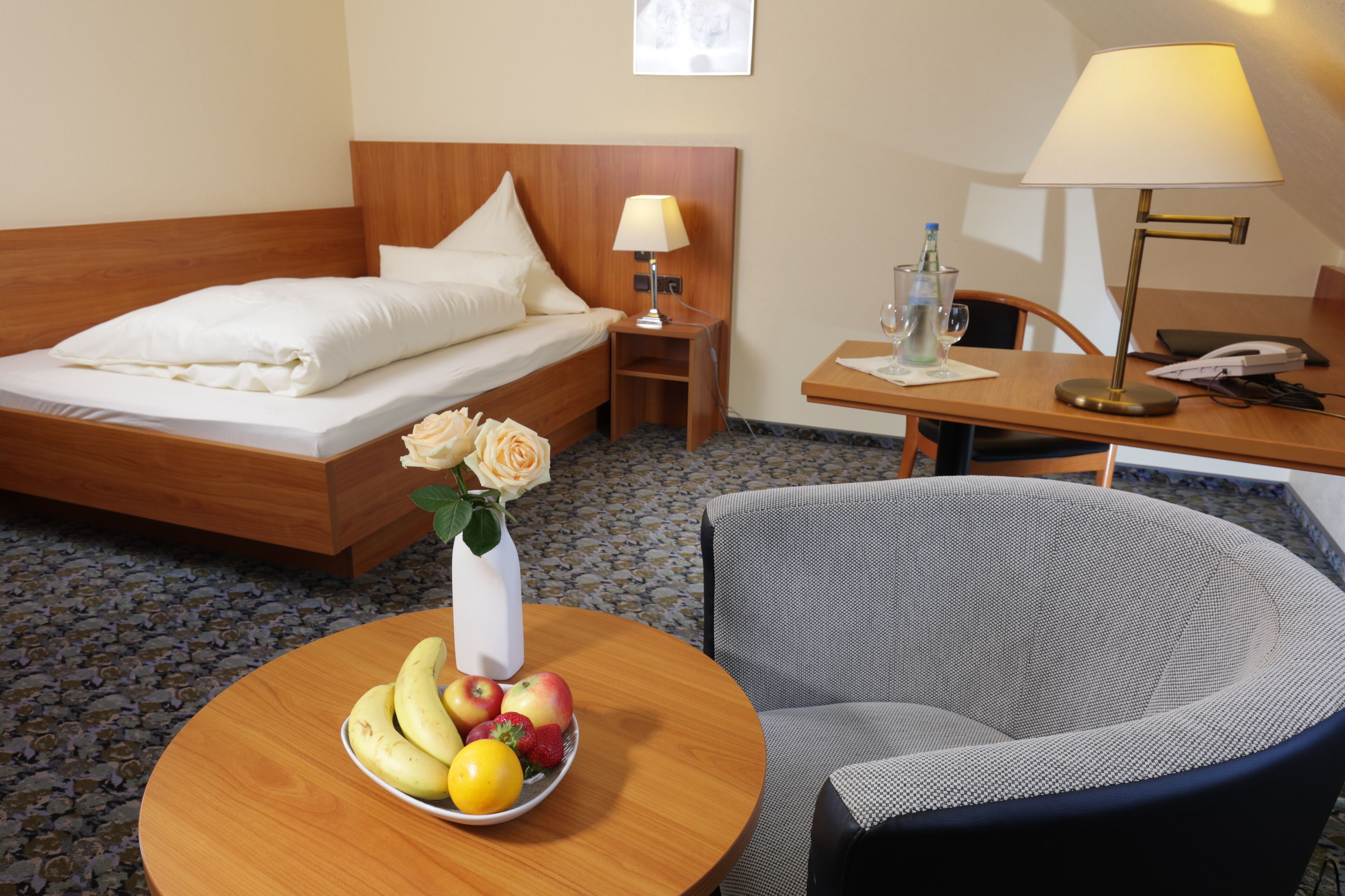 Quarto de hotel com cama, mesa redonda com frutas, uma cadeira, escrivaninha com abajur e pequena mesa com vaso de flores.