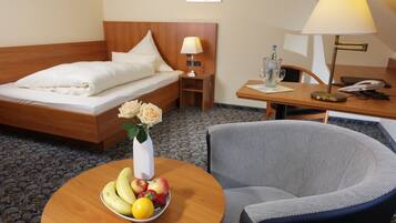 Quarto de hotel com cama, mesa redonda com frutas, uma cadeira, escrivaninha com abajur e pequena mesa com vaso de flores.
