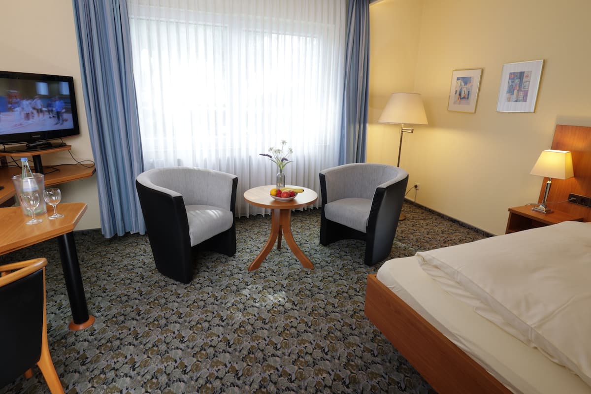 Quarto de hotel com cama, duas poltronas, uma mesinha redonda com um vaso de flores, televisão e luminária.