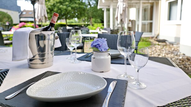 Mesa lista para comer al aire libre con botella de vino, copas y un florero con flores.