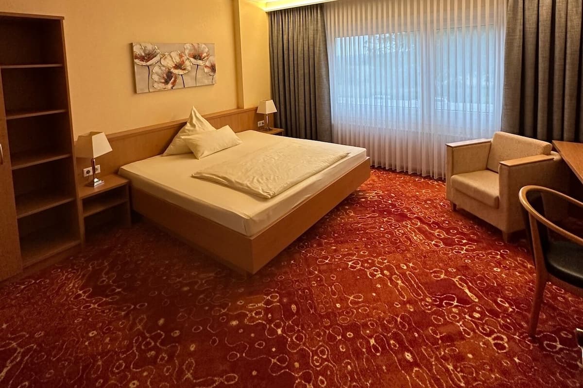Quarto de hotel com uma cama grande, uma escrivaninha, uma cadeira, uma estante de livros e um quadro na parede.