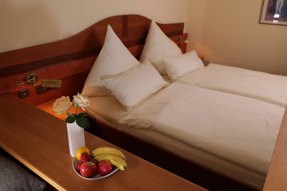 Quarto de hotel com cabeceira de madeira, cama com lençóis brancos, mesa de cabeceira com uma tigela de frutas e um vaso com flores.