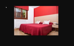 A bedroom with a red bedspread, a wooden headboard, and a window with a view of greenery.