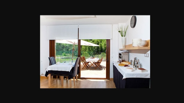 Een eetruimte met een tafel gedekt voor twee personen, uitzicht op de omgeving en een keuken met een klok aan de muur.