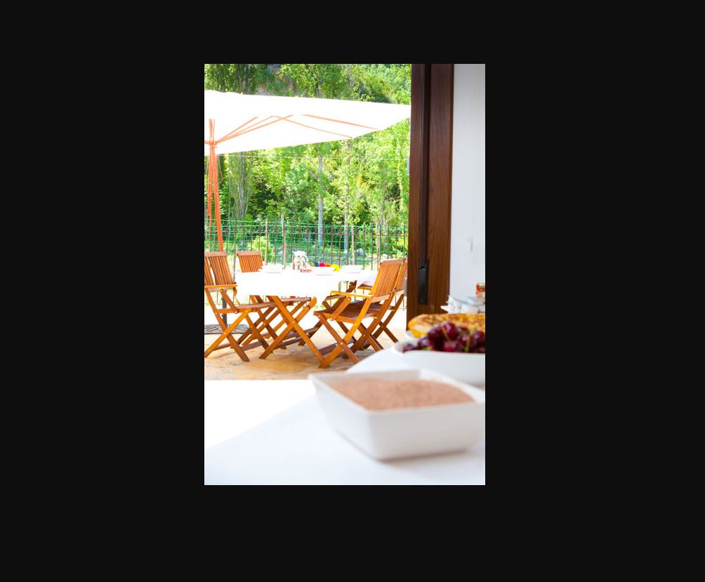 An outdoor seating area with wooden chairs and tables, a white umbrella, and a white table with a bowl of fruit.