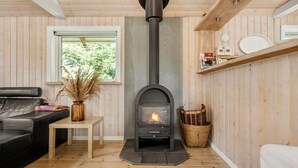 A cozy living room with a black wood-burning stove, a black leather sofa, a small wooden table with a vase of dried plants, and a window with a view of greenery. - Awesome home in Hovborg with WiFi (Hovborg)