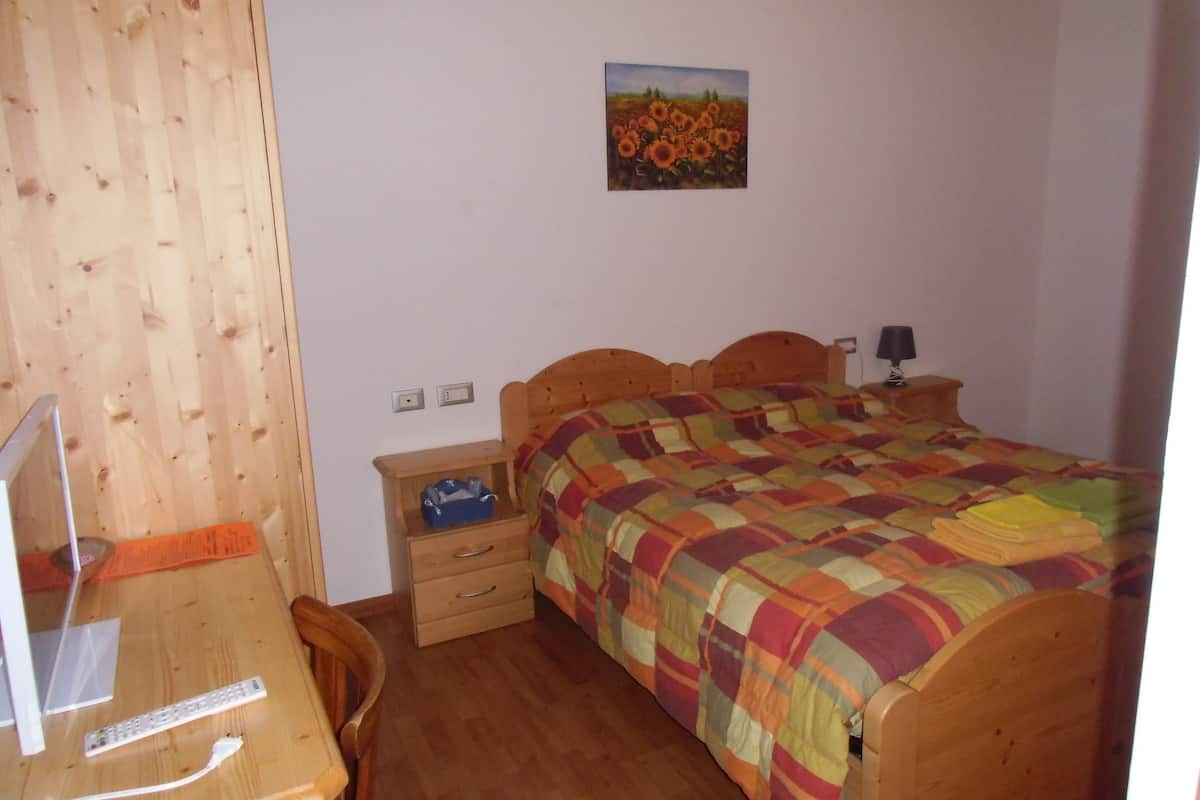 Un dormitorio con cama de madera, un escritorio con computadora, una silla, una mesita de noche con lámpara y un cuadro en la pared.