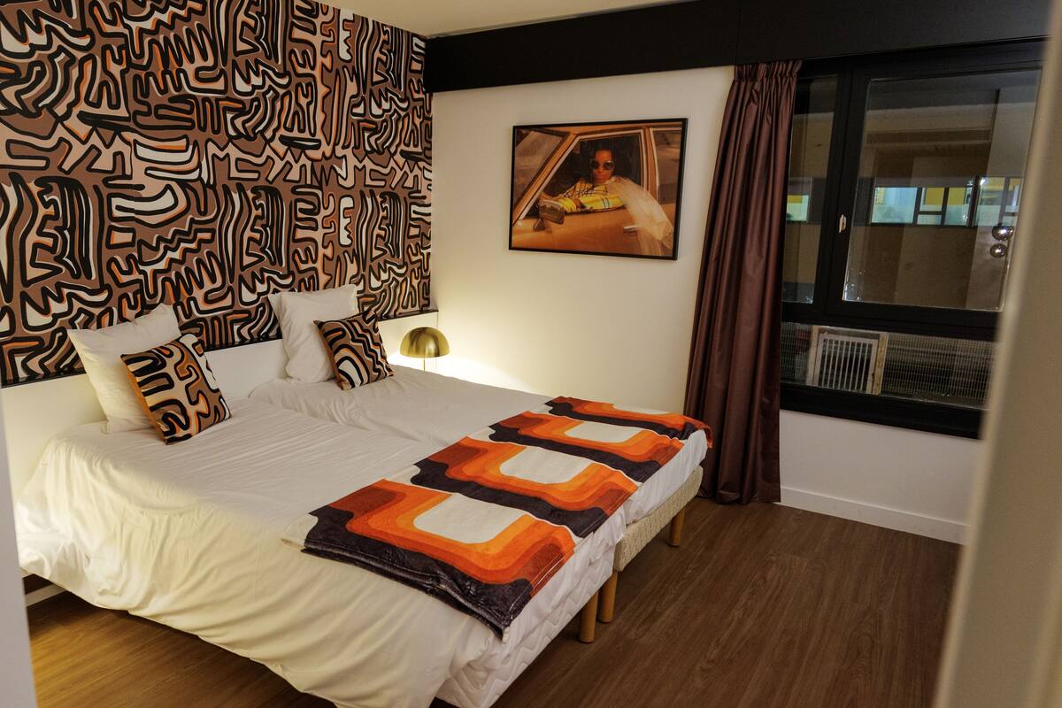 Un dormitorio moderno con un cabecero estampado, una cama grande con muchas almohadas, una manta decorativa y una ventana con cortinas.