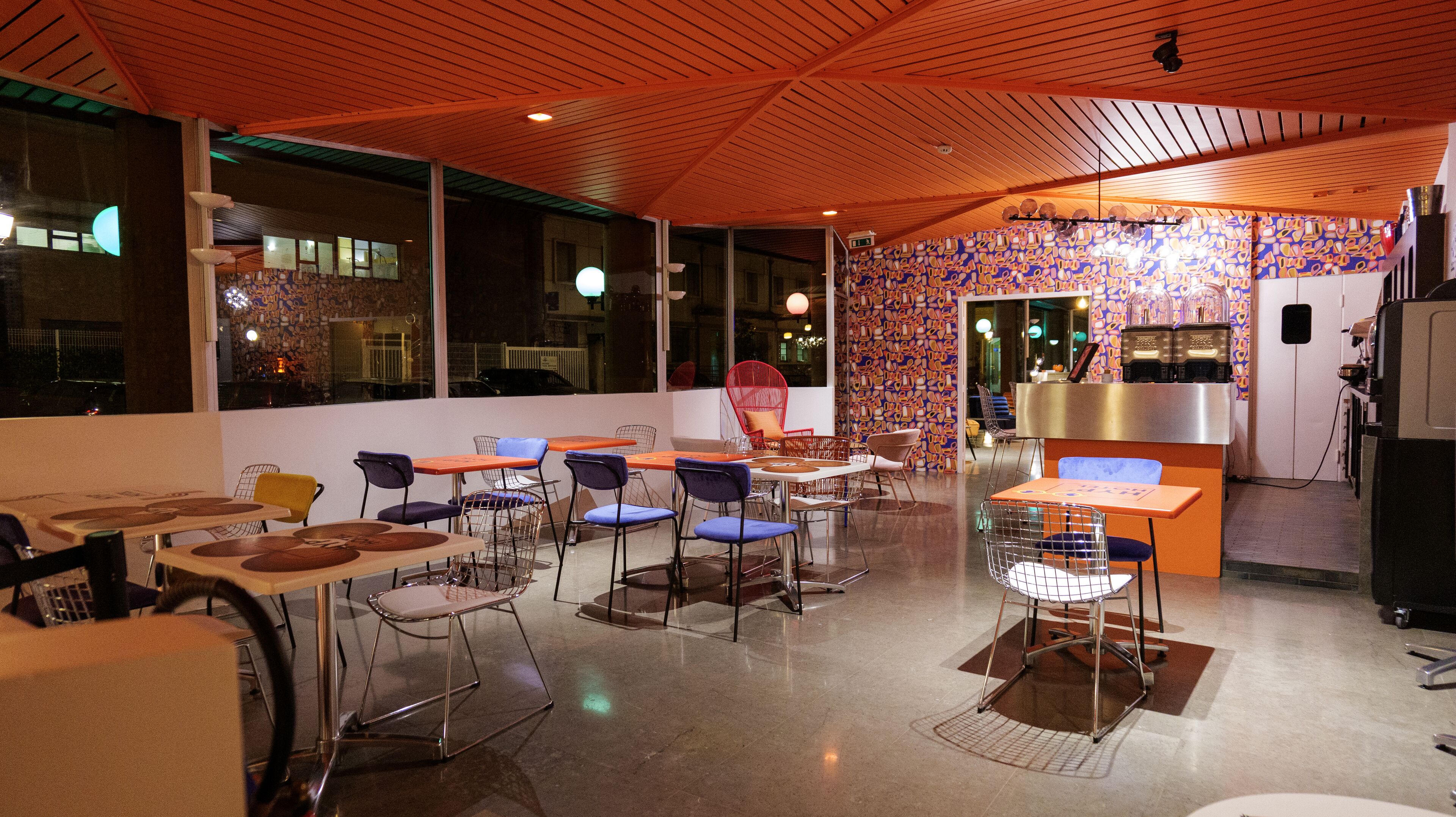 A modern restaurant interior with a variety of seating options, including tables and chairs, and a kitchen area visible in the background.
