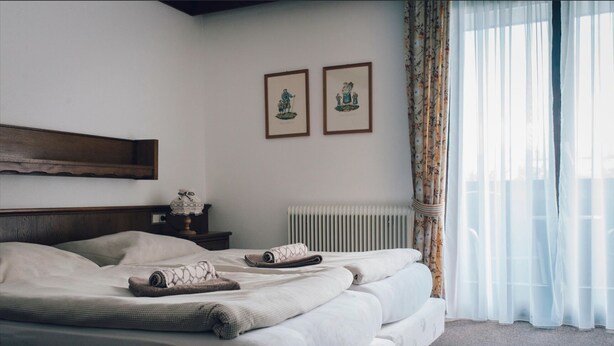 A neatly made bed with pillows and a blanket, a bedside table with a lamp, and a radiator. There are two framed pictures on the wall. - Pension Baranek (Kaprun)