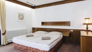 A double bed with a floral-patterned bed skirt, a wooden headboard, a bedside table with a lamp, and a framed picture on the wall. - Pension Baranek (Kaprun)