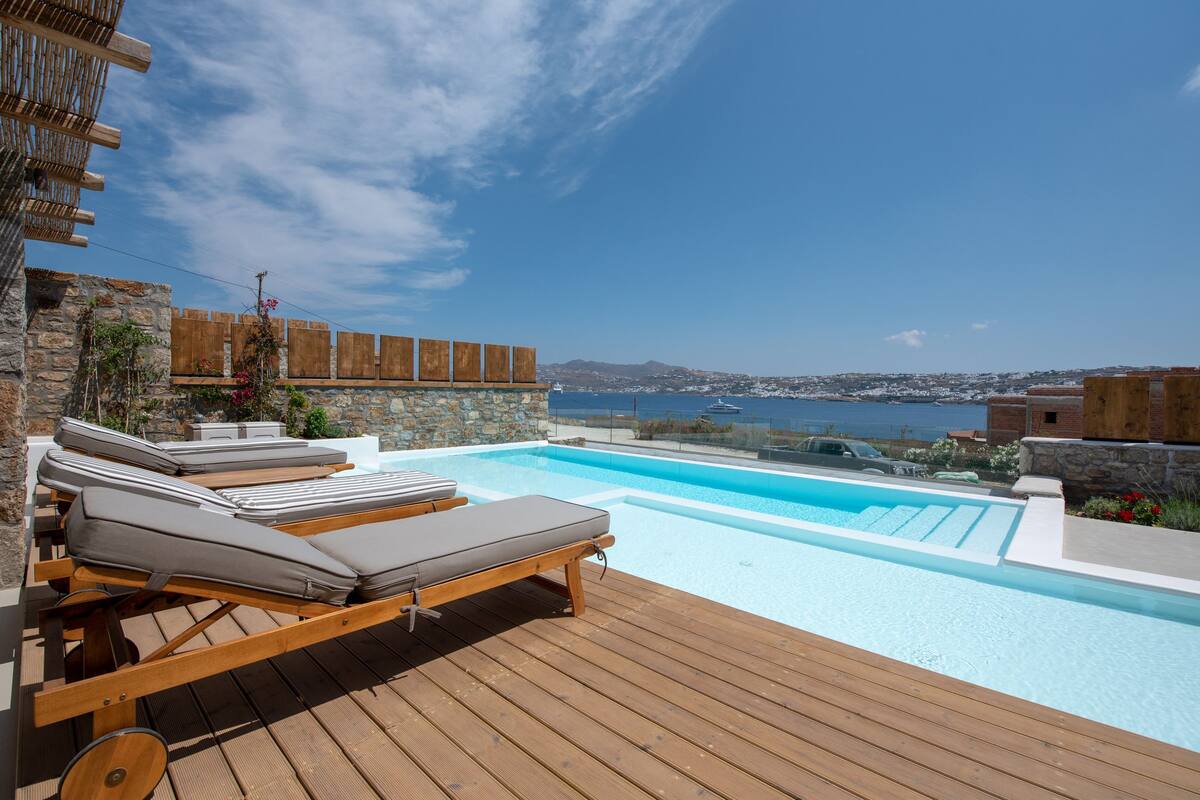 A wooden deck with lounge chairs and a swimming pool overlooking a coastal landscape.