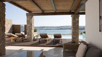 A covered outdoor seating area with stone pillars, lounge chairs, and a view of the sea.