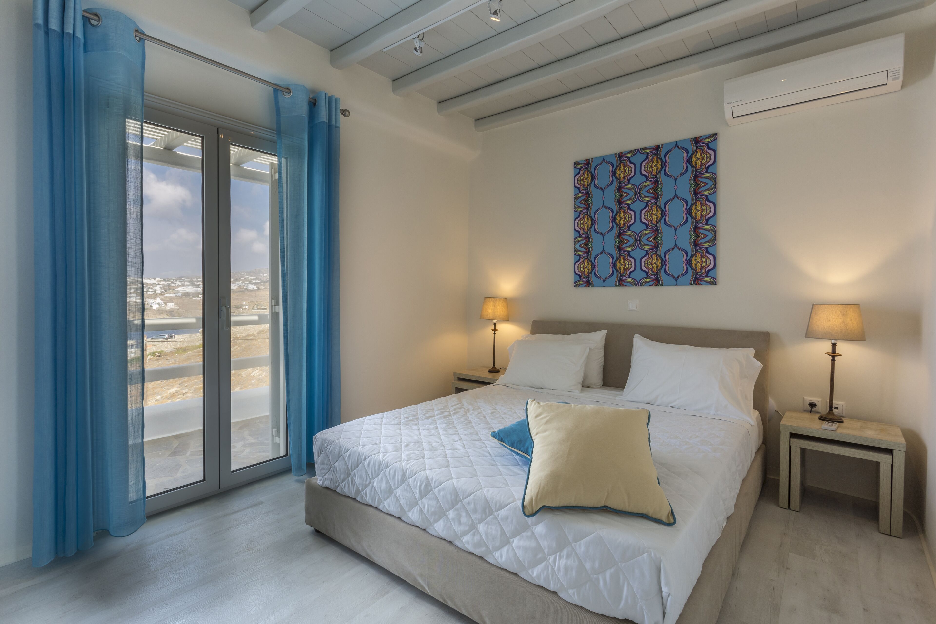 A bedroom with a large bed, two bedside tables with lamps, a window with blue curtains, and a wall-mounted air conditioner.