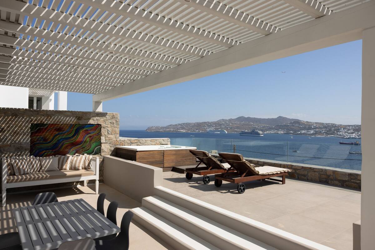A terrace with a sofa, lounge chairs, and a view of the sea and mountains.