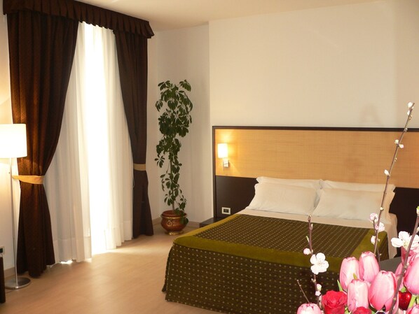 A hotel room with a bed, curtains, a lamp, a potted plant, and a vase with flowers.