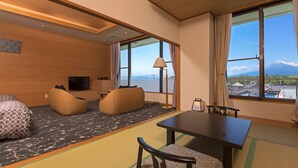 Ein Hotelzimmer mit Tatami-Boden, einem Esstisch, Stühlen, einer Couch, einem Fernseher und großen Fenstern mit Bergblick.