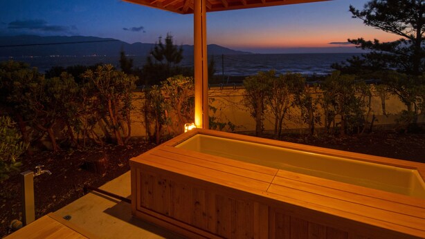 Ein hölzernes Whirlpool unter einer Holzpergola mit Blick auf das Meer und die Berge in der Abenddämmerung.