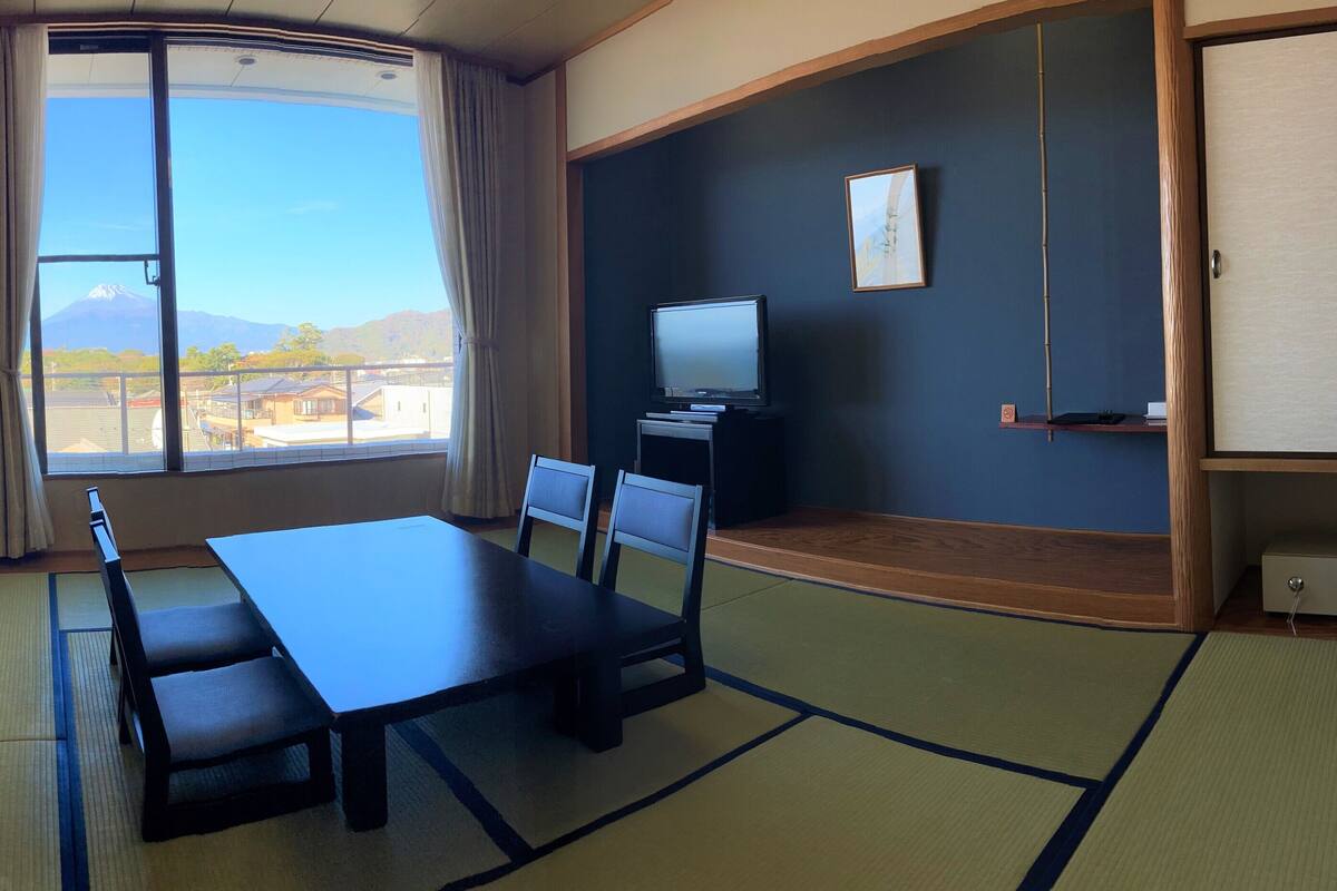 Una habitación con suelo de tatami, una mesa baja con sillas, un televisor sobre un soporte y vistas a la montaña a través de un ventanal grande.