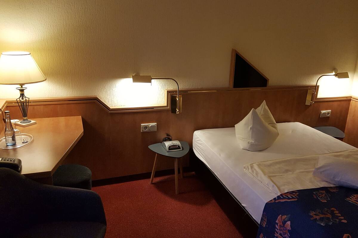 A hotel room with a bed, a desk, a chair, a small table, and two lamps.